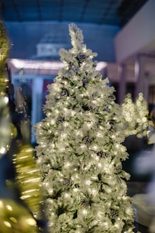 A beautifully decorated white artificial Christmas tree with glowing lights and ornamental pinecones standing in a dimly lit room. The bokeh effect creates a soft blur of lights in the background, enhancing a cozy and festive atmosphere.