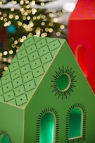 A brightly colored decorative model of a house with a green roof and black geometric patterns is in the foreground. The house features cut-out windows and circular designs. In the background, warm, blurred lights resembling bokeh effects suggest a festive or holiday setting.