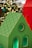 A brightly colored decorative model of a house with a green roof and black geometric patterns is in the foreground. The house features cut-out windows and circular designs. In the background, warm, blurred lights resembling bokeh effects suggest a festive or holiday setting.