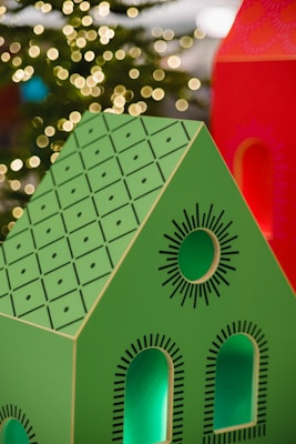 A brightly colored decorative model of a house with a green roof and black geometric patterns is in the foreground. The house features cut-out windows and circular designs. In the background, warm, blurred lights resembling bokeh effects suggest a festive or holiday setting.