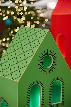 A brightly colored decorative model of a house with a green roof and black geometric patterns is in the foreground. The house features cut-out windows and circular designs. In the background, warm, blurred lights resembling bokeh effects suggest a festive or holiday setting.