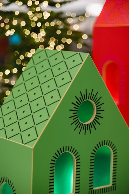 A brightly colored decorative model of a house with a green roof and black geometric patterns is in the foreground. The house features cut-out windows and circular designs. In the background, warm, blurred lights resembling bokeh effects suggest a festive or holiday setting.