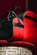 Two stylish bags with black and red leather accents are elegantly displayed on a patterned surface. The bags feature sleek designs with prominent black straps and zippers. The red sections have a glossy finish with a script-like embellishment.