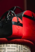 Two stylish bags with black and red leather accents are elegantly displayed on a patterned surface. The bags feature sleek designs with prominent black straps and zippers. The red sections have a glossy finish with a script-like embellishment.