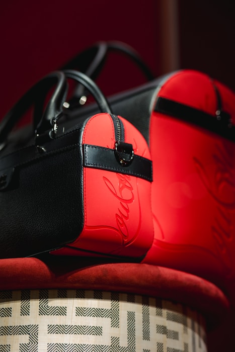 Two stylish bags with black and red leather accents are elegantly displayed on a patterned surface. The bags feature sleek designs with prominent black straps and zippers. The red sections have a glossy finish with a script-like embellishment.