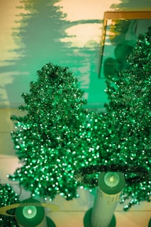 Sparkling green artificial trees decorated with vibrant green lights. The lighting casts soft green shadows on the wall, creating a festive and magical atmosphere. Two cylinder-shaped objects with halos are also visible at the bottom.