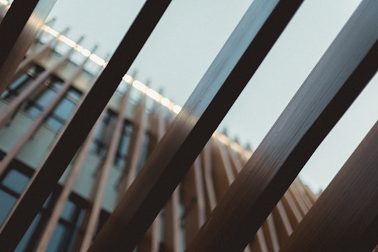 Wooden beams are arranged in parallel, drawing the eye toward a modern building facade with glass windows in the background. The beams are juxtaposed against a clear sky, creating a sense of depth and perspective.