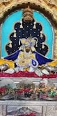 A richly adorned idol is placed in a decorative alcove, featuring elaborate garments in blue and yellow with intricate beadwork and embroidery. The setting includes ornate metallic elements such as a golden crown above, and silver statues of cows flanking the idol. The background is a combination of turquoise and black fabric adorned with tassels. Red roses are spread beneath the idol, alongside currency notes, and there are smaller altars with various figures and colorful decorations.