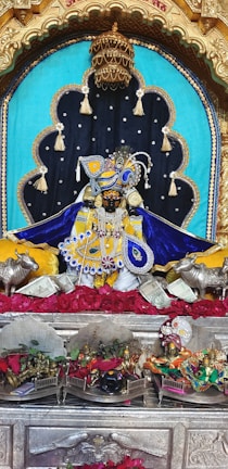 A richly adorned idol is placed in a decorative alcove, featuring elaborate garments in blue and yellow with intricate beadwork and embroidery. The setting includes ornate metallic elements such as a golden crown above, and silver statues of cows flanking the idol. The background is a combination of turquoise and black fabric adorned with tassels. Red roses are spread beneath the idol, alongside currency notes, and there are smaller altars with various figures and colorful decorations.