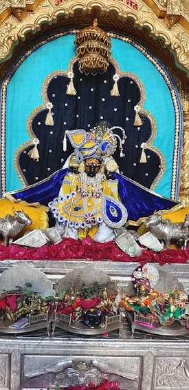 A richly adorned idol is placed in a decorative alcove, featuring elaborate garments in blue and yellow with intricate beadwork and embroidery. The setting includes ornate metallic elements such as a golden crown above, and silver statues of cows flanking the idol. The background is a combination of turquoise and black fabric adorned with tassels. Red roses are spread beneath the idol, alongside currency notes, and there are smaller altars with various figures and colorful decorations.