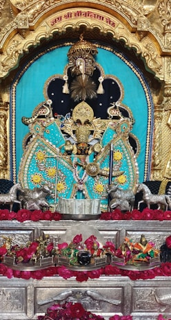 An ornate and colorful deity is depicted in traditional attire, featuring vibrant turquoise with gold and intricate embellishments. The idol is centered within a golden arched frame with detailed carvings. Surrounding the deity are decorative silver horse figures and a garland of red roses. The setup includes a silver platform with additional small gold-colored statues and elements symbolizing a religious setting.