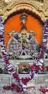 An ornate and colorful Hindu deity idol is centered in the image, set against a vibrant orange backdrop. The idol is adorned with intricate beadwork, jewels, and fabric. Surrounding the deity are silver animal figures on either side. A large garland made of red roses and currency notes decorates the idol. The setting features gold embellishments and a carved arch. At the base, there are offerings including flowers and possibly food items.