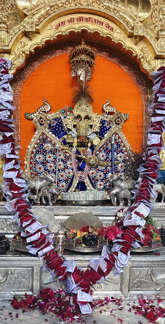 An ornate and colorful Hindu deity idol is centered in the image, set against a vibrant orange backdrop. The idol is adorned with intricate beadwork, jewels, and fabric. Surrounding the deity are silver animal figures on either side. A large garland made of red roses and currency notes decorates the idol. The setting features gold embellishments and a carved arch. At the base, there are offerings including flowers and possibly food items.