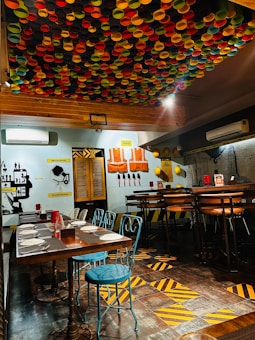 A vibrant restaurant interior with a ceiling covered in colorful caps, creating a lively atmosphere. The walls display industrial-themed decorations, including construction vests and tools. Wooden tables are set with placemats and chairs, some in blue metal and others in leather. The floor features a pattern of yellow and black stripes.