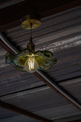 A vintage-style light bulb with visible filaments is encased in a textured, translucent glass fixture. The fixture is suspended from a metal ceiling with visible beams and corrugated roofing.