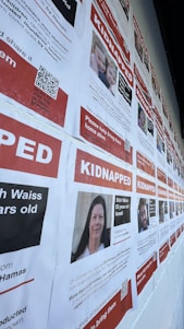 A wall covered with multiple posters that have a red and white color scheme, each with the word 'KIDNAPPED' prominently displayed at the top. The posters include photos and detailed information about several individuals, including their names and circumstances of their kidnapping. QR codes are present on each poster, possibly for more information or sharing purposes.