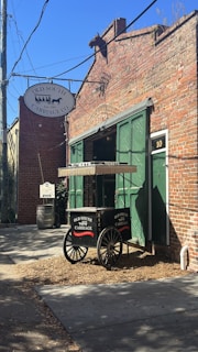 A brick building with a rustic style, featuring a large green double door and a decorative horse head protruding from the top of the building. A sign reading 'Old South Carriage Co.' is attached to the building. Below the sign, a two-wheeled cart with the same name displayed is positioned near the door. The area around the building includes a wooden barrel and a small patch of dirt.
