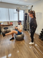 A man is balancing on an exercise ball while extending one leg forward. He is wearing athletic wear, including a cap, t-shirt, shorts, and sneakers. A woman stands nearby, observing him. The room contains gym equipment, including a cable machine and a set of dumbbells. The floor is wooden, and there are vertical blinds on the window.