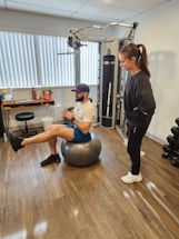 A man is balancing on an exercise ball while extending one leg forward. He is wearing athletic wear, including a cap, t-shirt, shorts, and sneakers. A woman stands nearby, observing him. The room contains gym equipment, including a cable machine and a set of dumbbells. The floor is wooden, and there are vertical blinds on the window.