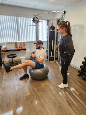A man is balancing on an exercise ball while extending one leg forward. He is wearing athletic wear, including a cap, t-shirt, shorts, and sneakers. A woman stands nearby, observing him. The room contains gym equipment, including a cable machine and a set of dumbbells. The floor is wooden, and there are vertical blinds on the window.