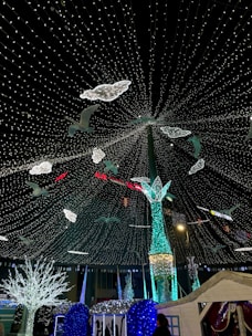 A dazzling display of decorative lights arranged to create a festive atmosphere. Birds and clouds made of bright lights are suspended in the air, creating a whimsical and magical scene. Below, luminous trees and a large, ornate light structure add to the enchanting setting.