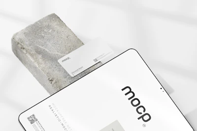 A business card with the text 'mocp' and 'Personal Brand' is placed on a rectangular concrete block. Below it, a tablet or large device displays 'mocp' in bold lettering and some text and QR codes. The setup is arranged on a white background with soft shadows.