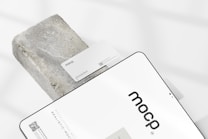 A business card with the text 'mocp' and 'Personal Brand' is placed on a rectangular concrete block. Below it, a tablet or large device displays 'mocp' in bold lettering and some text and QR codes. The setup is arranged on a white background with soft shadows.