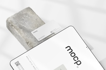 A business card with the text 'mocp' and 'Personal Brand' is placed on a rectangular concrete block. Below it, a tablet or large device displays 'mocp' in bold lettering and some text and QR codes. The setup is arranged on a white background with soft shadows.