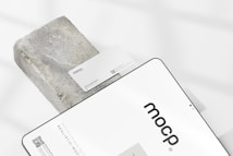 A business card with the text 'mocp' and 'Personal Brand' is placed on a rectangular concrete block. Below it, a tablet or large device displays 'mocp' in bold lettering and some text and QR codes. The setup is arranged on a white background with soft shadows.