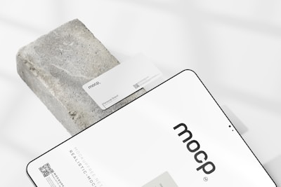 A business card with the text 'mocp' and 'Personal Brand' is placed on a rectangular concrete block. Below it, a tablet or large device displays 'mocp' in bold lettering and some text and QR codes. The setup is arranged on a white background with soft shadows.