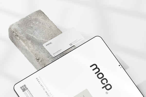 A business card with the text 'mocp' and 'Personal Brand' is placed on a rectangular concrete block. Below it, a tablet or large device displays 'mocp' in bold lettering and some text and QR codes. The setup is arranged on a white background with soft shadows.