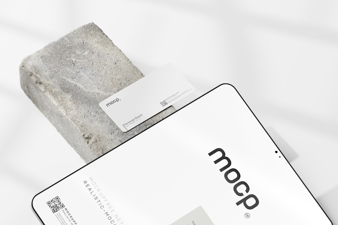 A business card with the text 'mocp' and 'Personal Brand' is placed on a rectangular concrete block. Below it, a tablet or large device displays 'mocp' in bold lettering and some text and QR codes. The setup is arranged on a white background with soft shadows.