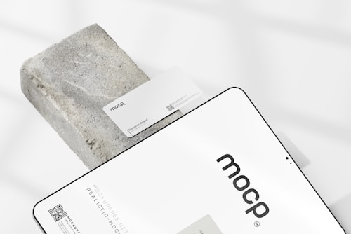 A business card with the text 'mocp' and 'Personal Brand' is placed on a rectangular concrete block. Below it, a tablet or large device displays 'mocp' in bold lettering and some text and QR codes. The setup is arranged on a white background with soft shadows.