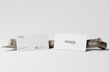 Two white business cards are displayed with a simple design featuring the text 'mocp' on each. They are positioned against a plain background and rest on a piece of driftwood.