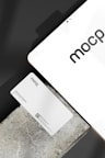 A business card with 'mocp' branding placed on a concrete surface, accompanied by a modern electronic device displaying the same text. The design is minimalist, featuring white and gray tones. A QR code is present on the card alongside the text 'Personal Brand'.