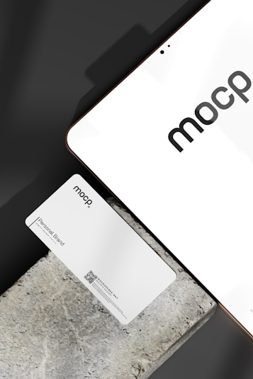 A business card with 'mocp' branding placed on a concrete surface, accompanied by a modern electronic device displaying the same text. The design is minimalist, featuring white and gray tones. A QR code is present on the card alongside the text 'Personal Brand'.