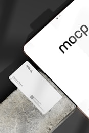 A business card with 'mocp' branding placed on a concrete surface, accompanied by a modern electronic device displaying the same text. The design is minimalist, featuring white and gray tones. A QR code is present on the card alongside the text 'Personal Brand'.