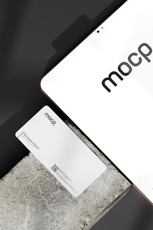 A business card with 'mocp' branding placed on a concrete surface, accompanied by a modern electronic device displaying the same text. The design is minimalist, featuring white and gray tones. A QR code is present on the card alongside the text 'Personal Brand'.