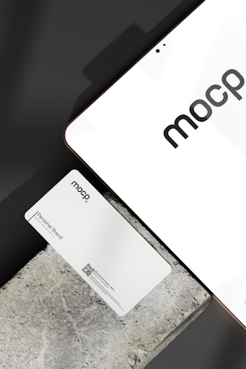 A business card with 'mocp' branding placed on a concrete surface, accompanied by a modern electronic device displaying the same text. The design is minimalist, featuring white and gray tones. A QR code is present on the card alongside the text 'Personal Brand'.