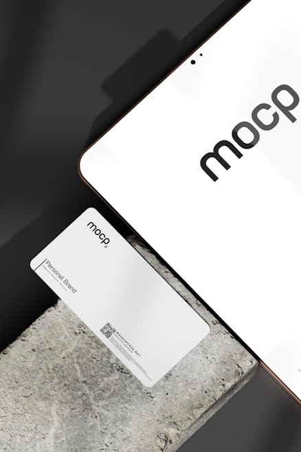 A business card with 'mocp' branding placed on a concrete surface, accompanied by a modern electronic device displaying the same text. The design is minimalist, featuring white and gray tones. A QR code is present on the card alongside the text 'Personal Brand'.