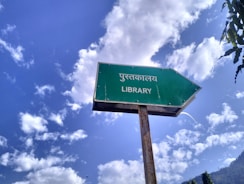 A weathered green sign with an arrow pointing right and the text in Hindi and English for 'Library'. The background features a bright blue sky with scattered white clouds, creating a serene and pleasant atmosphere. Some trees or foliage can be seen at the edges.