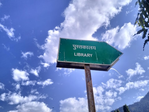 A weathered green sign with an arrow pointing right and the text in Hindi and English for 'Library'. The background features a bright blue sky with scattered white clouds, creating a serene and pleasant atmosphere. Some trees or foliage can be seen at the edges.