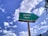 A weathered green sign with an arrow pointing right and the text in Hindi and English for 'Library'. The background features a bright blue sky with scattered white clouds, creating a serene and pleasant atmosphere. Some trees or foliage can be seen at the edges.