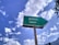 A weathered green sign with an arrow pointing right and the text in Hindi and English for 'Library'. The background features a bright blue sky with scattered white clouds, creating a serene and pleasant atmosphere. Some trees or foliage can be seen at the edges.