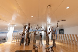 An interior space features wooden floorboards and a modern aesthetic, with several tree-like sculptures extending from the floor to the ceiling. Three people are walking through the space, with blurry motion suggesting movement. Shelving units occupy the background, and the ceiling is reflective, enhancing the spacious feel.