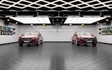 A modern garage with two identical red cars parked side by side. The ceiling features geometric, grid-like lighting that is bright and reflective. The walls are decorated with colorful car detailing supplies. The floor has a textured, dark tile surface.