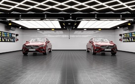 A modern garage with two identical red cars parked side by side. The ceiling features geometric, grid-like lighting that is bright and reflective. The walls are decorated with colorful car detailing supplies. The floor has a textured, dark tile surface.