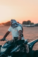 A person wearing a white t-shirt and a headscarf is seated on a quad bike in a desert setting, with the sun setting in the background casting a warm glow.
