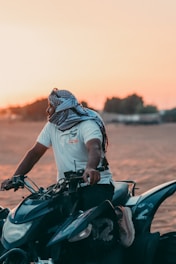 A person wearing a white t-shirt and a headscarf is seated on a quad bike in a desert setting, with the sun setting in the background casting a warm glow.