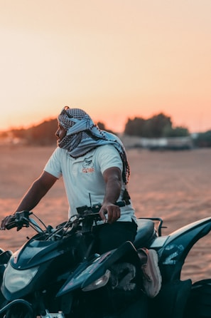 A person wearing a white t-shirt and a headscarf is seated on a quad bike in a desert setting, with the sun setting in the background casting a warm glow.